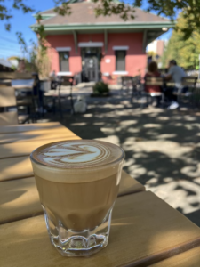 Closeup of a cortado in a glass, sitting on a wooden table. A brick building is in the background.