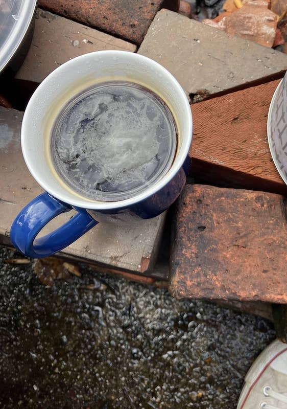 A moody photo looking down on a mug of black coffee atop a pile of bricks, with at the tip of a shoe in the lower right corner