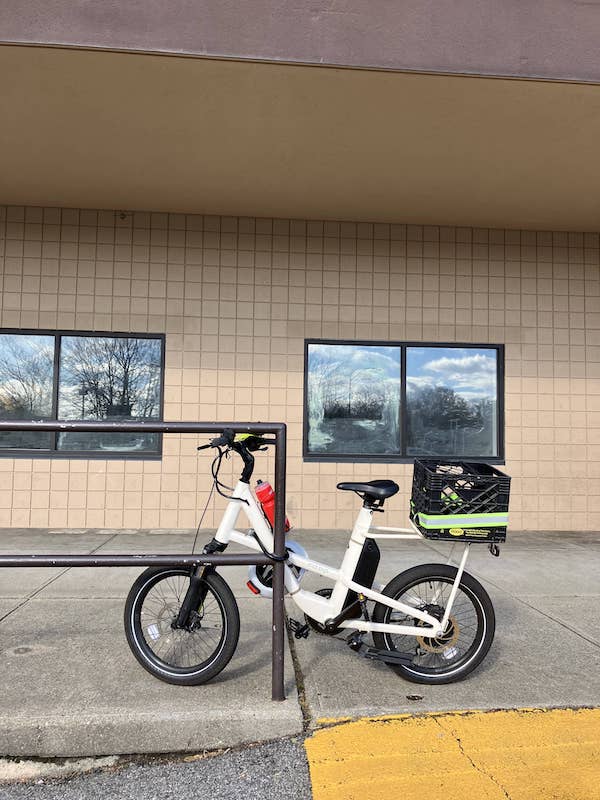 A white cargo e-bike with a milk crate on the back is locked to a railing next to an industrial-looking building