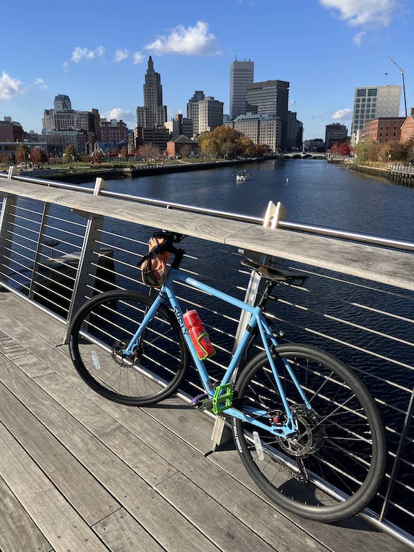 A light-blue bike rests against a railing on a bridge with river below and the city Providence skyline in the background
