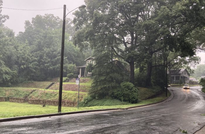 Photo of a wet road going down a hill next to a beautiful terraced lawn. A tiny bus stop sign stands next to a power pole.