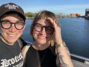 Photo of Erin and their wife Courtnie standing in front of the Providence skyline. Erin is wearing a black Duke's mayo cap and a black t-shirt. Courtnie is wearing a black tank top. They are both smiling big!
